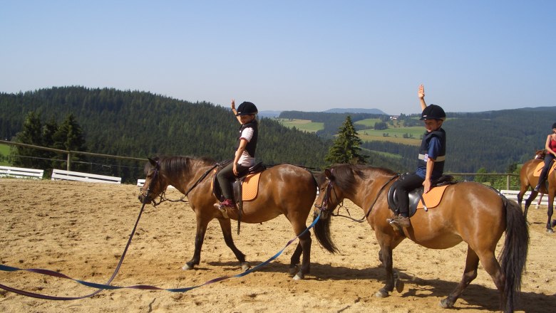 Kinder reiten auf Ponys in einer ländlichen Umgebung mit Hügeln im Hintergrund.