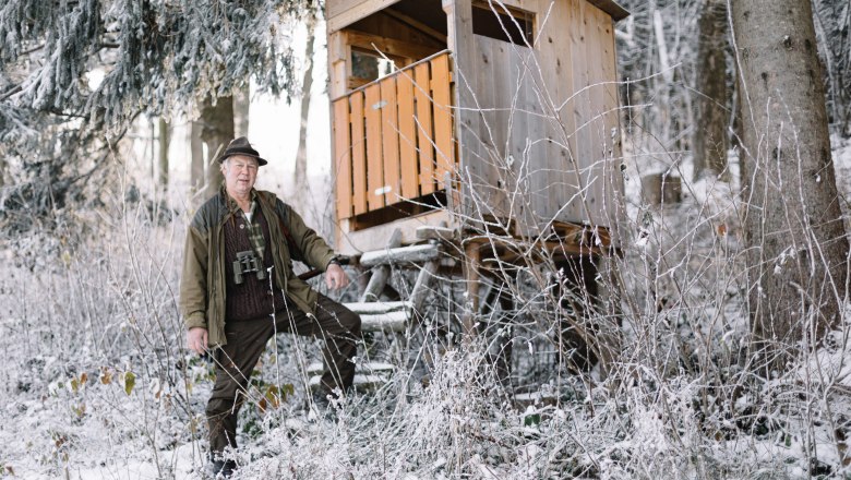 Ein Mann in Jagdkleidung steht neben einem Hochsitz im verschneiten Wald.