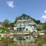 Ein gro&szlig;es, gelbes Haus mit blauen Akzenten spiegelt sich in einem Teich. Im Vordergrund sind gr&uuml;ne Liegest&uuml;hle und B&auml;ume zu sehen.