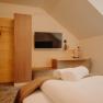 Ein gem&uuml;tliches Hotelzimmer fotografiert vom Bett aus. Gegen&uuml;ber vom Bett h&auml;ngt ein Flachbildfernsehener an der Wand. Daneben ist eine Garderobe aus hellem Holz, die Wand ist auch zum Teil mit Holz verkleidet. Dort befindet sich auch eine Ablagefl&auml;che und ein Schreibtisch mit Sessel und Lampe. Das Zimmer hat eine Dachschr&auml;ge.