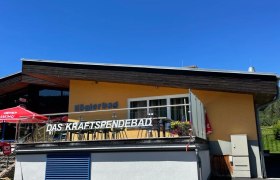 Gelbes Gebäude mit der Aufschrift 'Köglerbad Das Kraftspendebad' und Sonnenschirmen auf der Terrasse.