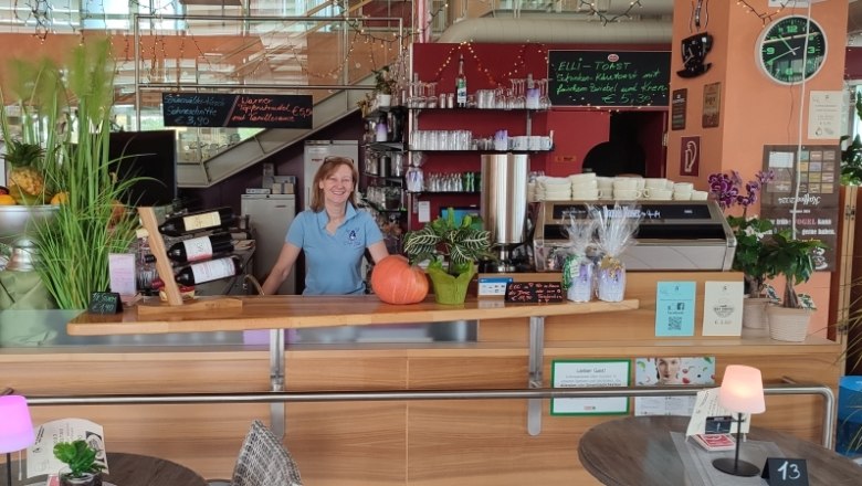 Eine Frau steht hinter der Theke eines Caf&eacute;s mit Pflanzen, Weinflaschen und einer Kaffeemaschine.