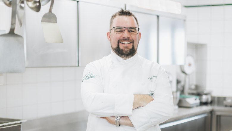 Egy mosolygó, fehér egyenruhás szakács áll egy profi konyhában.