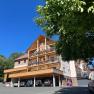 Hotelgebäude mit Holzbalkonen und Carport, umgeben von Bäumen, unter blauem Himmel.