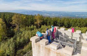 Drei Wanderer auf einem Aussichtsturm mit Blick auf bewaldete Hügel.