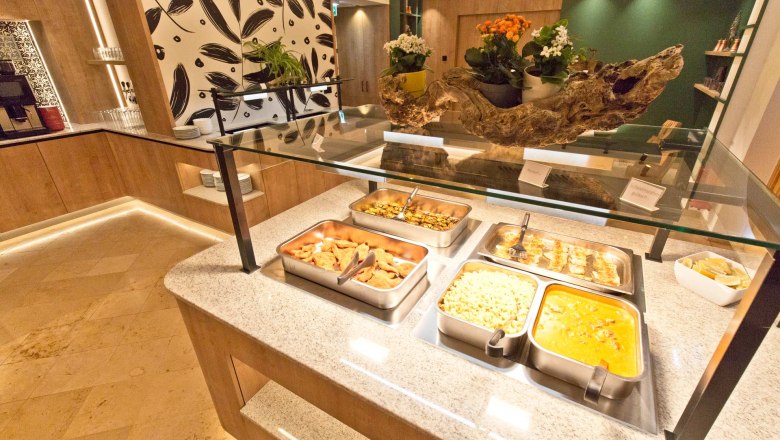 Buffetbereich mit verschiedenen Speisen in Edelstahlbehältern, dekoriert mit Pflanzen und Holzelementen.