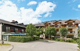Ein traditionelles Gasthaus mit Holzbalkonen und Blumen, umgeben von Bäumen und Bergen unter blauem Himmel.