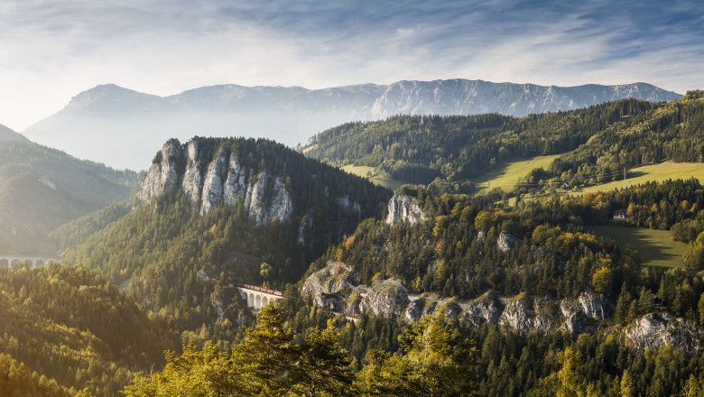 Panoramablick auf die Semmeringbahn in einer bergigen Landschaft mit Wäldern und einem Viadukt.