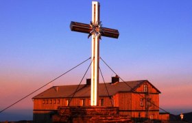 Großes Gipfelkreuz vor dem Alois-Günther-Haus bei Sonnenuntergang.