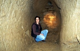 Junge Frau hockt in einem schmalen, beleuchteten Tunnel aus Erde und Stein.