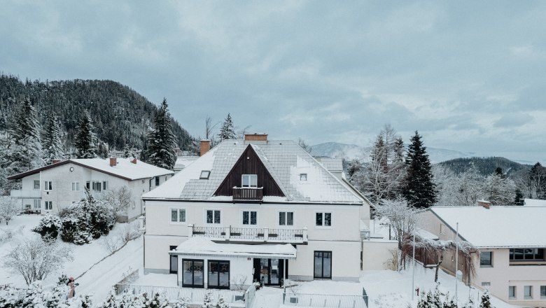 Ein gro&szlig;es, schneebedecktes Haus in einer winterlichen Landschaft mit Bergen im Hintergrund.