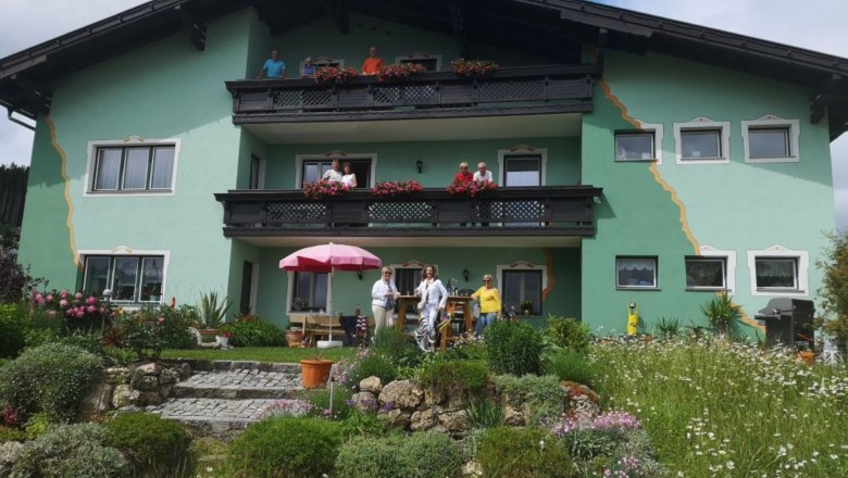 Ein grünes Haus mit Balkonen und Menschen davor, umgeben von einem blühenden Garten.