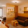 Modernes Hotelappartement mit Doppelbett, hellen Holzm&ouml;beln, ausziehbarer Couch und Raumtrenner aus Holz. Die Wand ist zum Teil mit Holz verkleidet und in der Wand sind Nischen, die in einem erdigen Gold gestrichen sind.