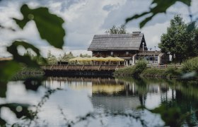 Ein rustikales Holzhaus mit gelben Sonnenschirmen spiegelt sich in einem ruhigen See, umgeben von grüner Natur.