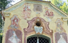 Fassade einer Kapelle mit religiösen Fresken und Inschrift.