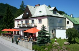 Ein zweistöckiges Gebäude mit der Aufschrift 'Löffler Pension-Café-Restaurant', umgeben von Bäumen und Blumen, mit orangenen Sonnenschirmen auf der Terrasse.
