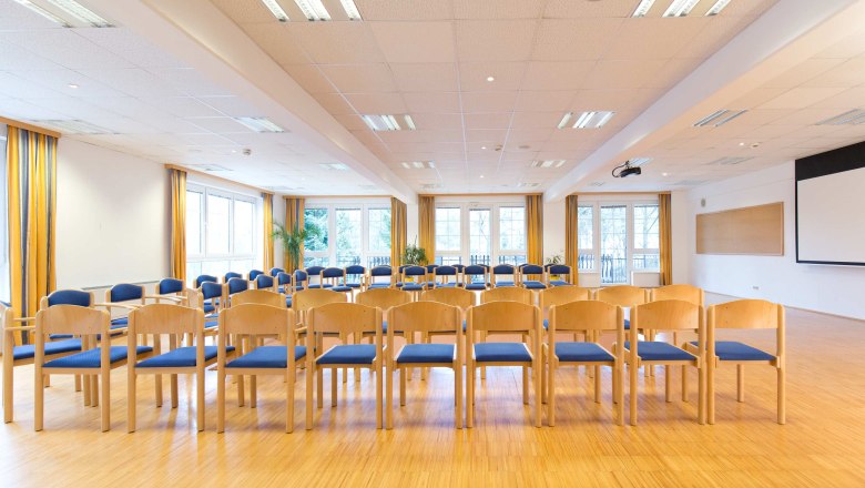 Ein heller Seminarraum mit Stuhlreihen, gro&szlig;en Fenstern und einem Projektor an der Decke.