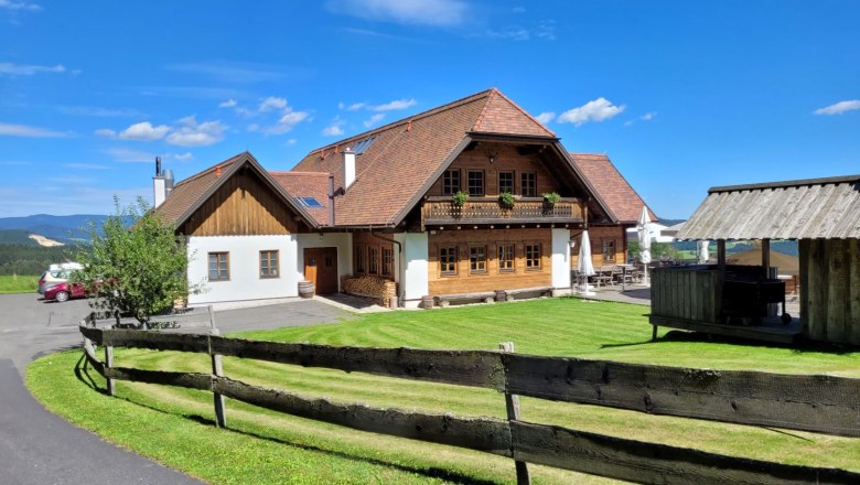 Traditionelles Holzhaus mit rotem Dach in ländlicher Umgebung, blauer Himmel, grüner Rasen, Holzzaun im Vordergrund.