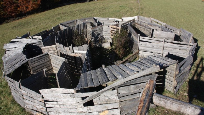 Ein aus Holzplanken gebautes Labyrinth auf einer Wiese, umgeben von B&auml;umen im Herbst.