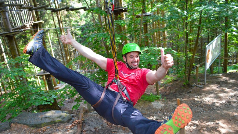 Person mit Helm und Sicherheitsgurt auf einer Seilrutsche im Wald.
