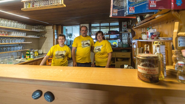 Drei Personen in gelben T-Shirts stehen hinter einer Bar in einem rustikalen Raum.