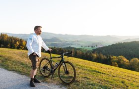 Ein Mann in weißem Hemd steht mit einem Fahrrad auf einem Hügel mit Blick auf eine grüne Landschaft.