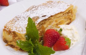Ein Stück Apfelstrudel mit Puderzucker, Erdbeeren und Schlagobers auf einem Teller.