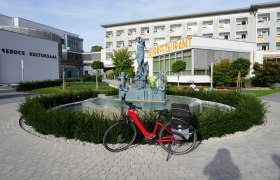 Brunnen mit einer Statue vor einem Geb&auml;ude mit der Aufschrift "Kurrestaurant", davor steht ein rotes Fahrrad..