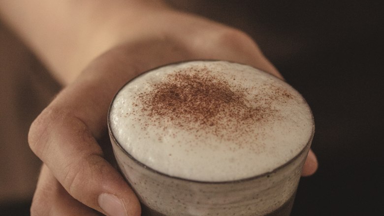 Nahaufnahme einer Hand, die ein Glas mit Cappuccino und Milchschaum hält.
