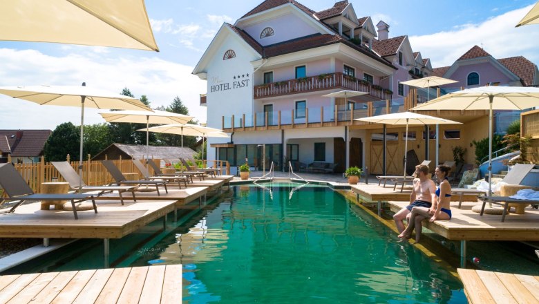 Mein Hotel Fast, © Karl Schrotter Außenansicht eines Hotels mit Naturpool und Sonnenschirmen im Vordergrund.
