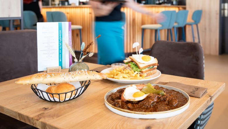 Ein Tisch in einem Restaurant mit regionalen Speisen: ein Teller mit Gulasch und Spiegelei, ein Sandwich mit Pommes und ein Brotkorb.