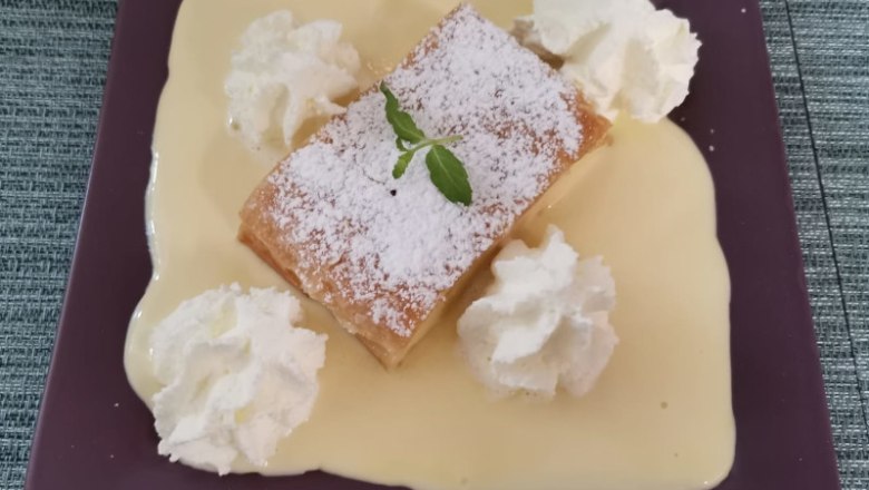 Ein Dessert auf einem lila Teller: Ein St&uuml;ck Topfenstrudel mit Puderzucker, umgeben von Vanilleso&szlig;e und Sahneh&auml;ubchen, garniert mit einem Minzblatt.