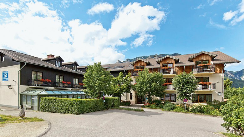 Ein traditionelles Gasthaus mit Holzbalkonen und Blumen, umgeben von Bäumen und Bergen unter blauem Himmel.
