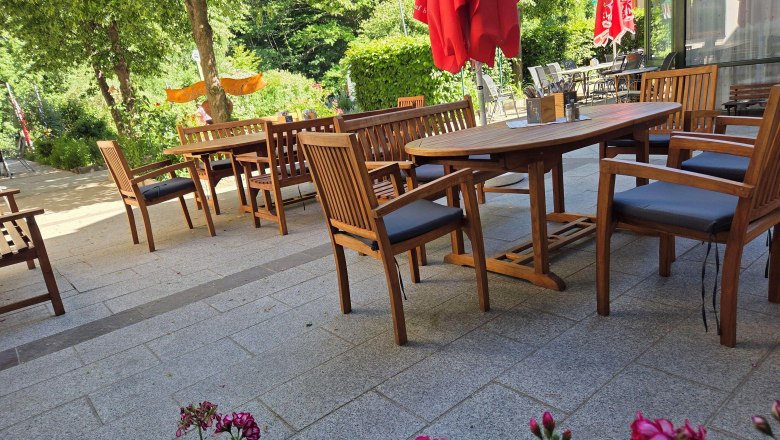 Ein gem&uuml;tlicher Gastgarten mit Holzm&ouml;beln und roten Sonnenschirmen, umgeben von gr&uuml;nen B&auml;umen und Blumen.