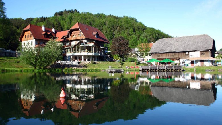 Ein Gasthof am See mit rotem Dach und Terrasse, umgeben von Bäumen, spiegelt sich im Teich davor wider.