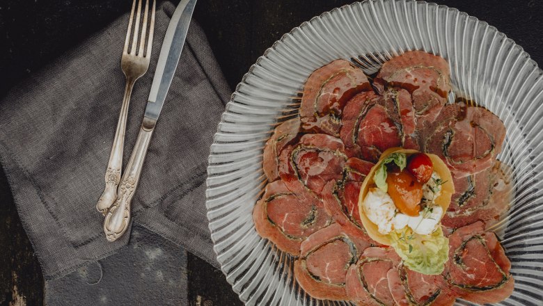 Ein Teller mit dünn geschnittenem Fleisch und einer kleinen Salatgarnitur in der Mitte, daneben Besteck auf einer Serviette.