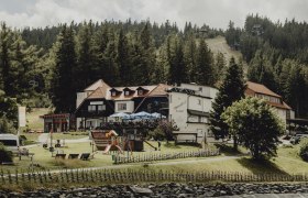 Ein Gasthaus in einer bergigen Waldlandschaft mit Spielplatz und Seilbahn im Hintergrund.
