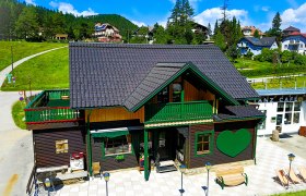 Ein traditionelles Holzhaus mit gr&uuml;nem Herzmotiv und Terrasse im Sommer, umgeben von gr&uuml;nen H&uuml;geln und blauem Himmel.