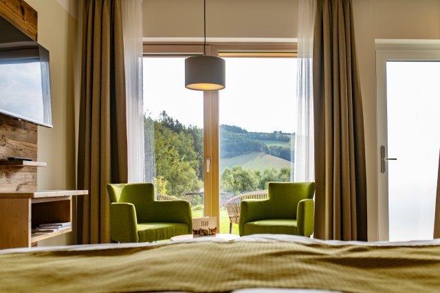 Innenansicht eines Gästezimmers mit zwei grünen Sesseln vor einem großen Fenster mit Blick auf eine hügelige Landschaft.