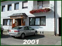 Einstöckiges Gebäude mit weißer Fassade und roten Blumenkästchen am Balkon.