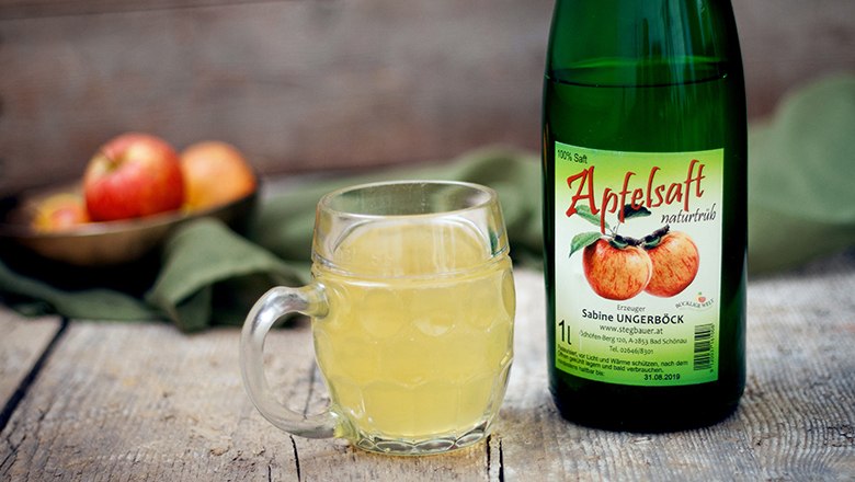 Eine Flasche Apfelsaft und ein Glas gefüllt mit Apfelsaft auf einem Holztisch, im Hintergrund ein grünes Tuch.