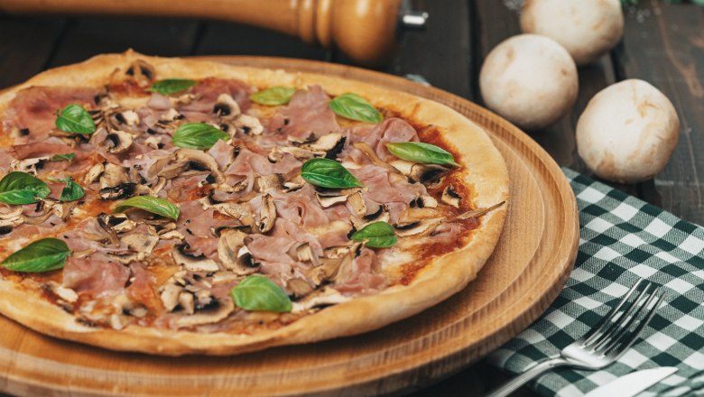 Pizza mit Schinken, Pilzen und Basilikum auf einem Holzbrett.