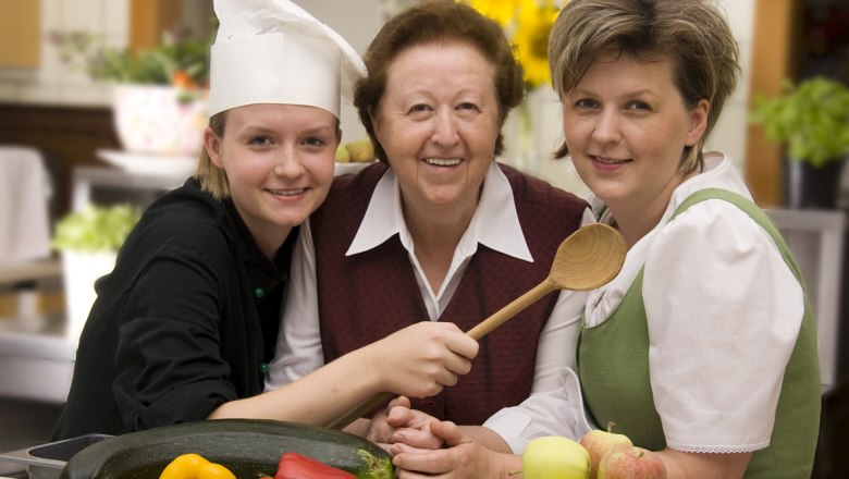 Drei Frauen in einer Küche, eine trägt eine Kochmütze, Gemüse und Obst im Vordergrund.