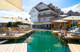 Außenansicht eines Hotels mit Naturpool und Sonnenschirmen im Vordergrund.