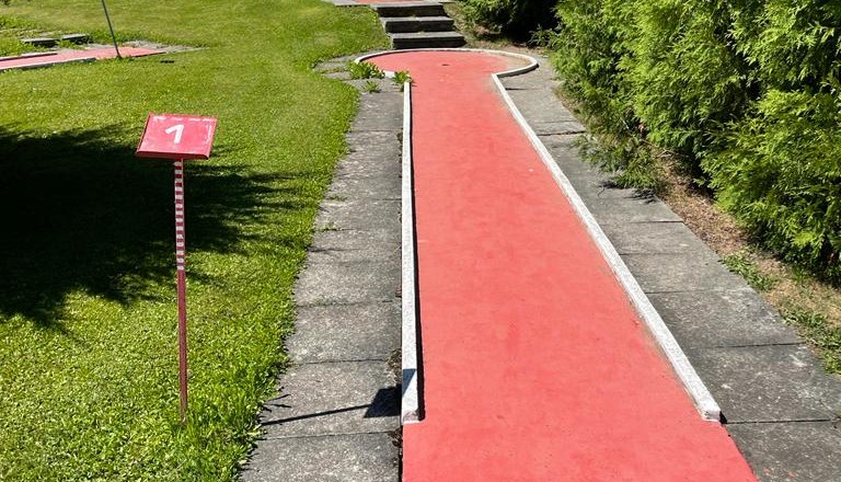 Minigolfbahn mit rotem Belag und grüner Hecke.