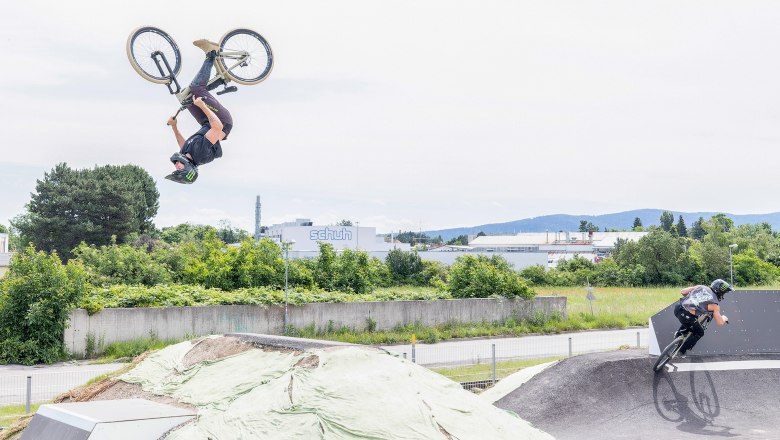Zwei BMX-Fahrer auf einer Pumptrack-Anlage, einer macht einen R&uuml;ckw&auml;rtssalto, der andere f&auml;hrt eine Kurve.