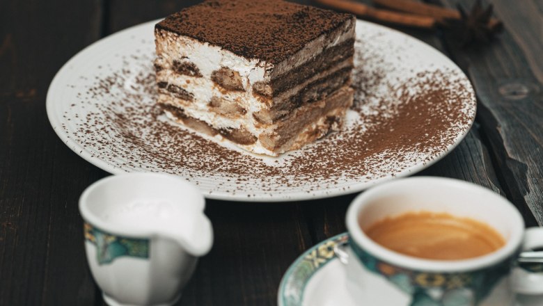 Ein Teller mit einem Stück Tiramisu, daneben eine Tasse Kaffee und ein Milchkännchen auf einem Holztisch.