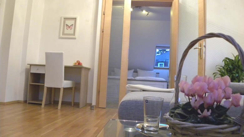 Innenraum mit Schreibtisch, Stuhl, Blumen im Korb und Blick ins Schlafzimmer.