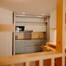 Moderne K&uuml;chenzeile in einem Apartment in hellen Farbt&ouml;nen. Man sieht eine Sp&uuml;le, eine Mikrowelle, eine gro&szlig;e Herdplatte und einen Wasserkocher. Die K&uuml;che kann hinter einer verschiebbaren Trennwand versteckt werden. Vor der K&uuml;che sieht man einen Tisch an der Wand und 4 Sessel.