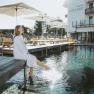 Frau in Bademantel sitzt am Rand des Naturpools eines Hotels mit Sonnenschirmen und Liegest&uuml;hlen.